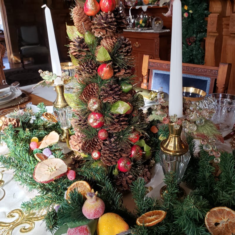 A pinecone and pomegranate centerpiece stands between two white candles on a festive table, with a wooden interior and a decorated tree in the background.