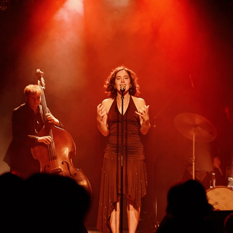 A female singer in a shimmering dress performs center stage under red lights, with a double bassist to her left and a drum kit in the background.