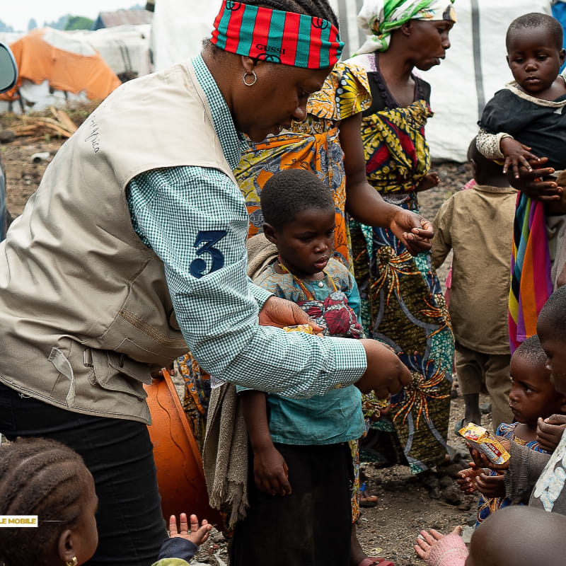 A woman in a tan vest and colorful headband hands out supplies to children and women in a camp with tents in the background.
