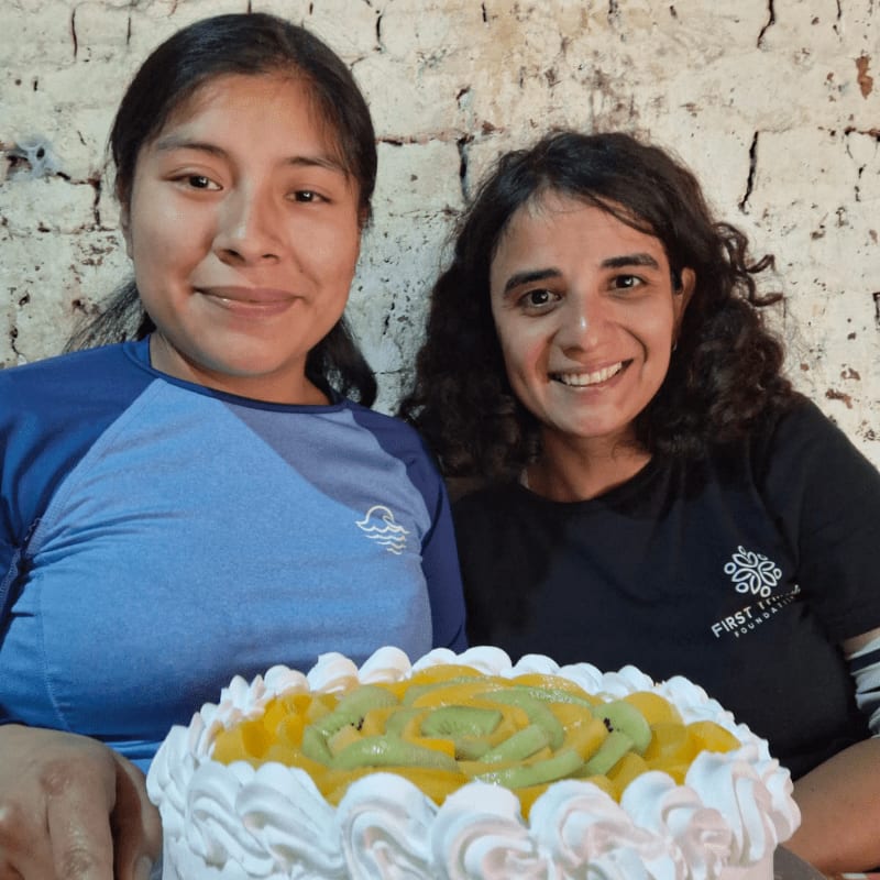Two smiling women pose behind a fruit-topped cake against a rustic white brick wall.