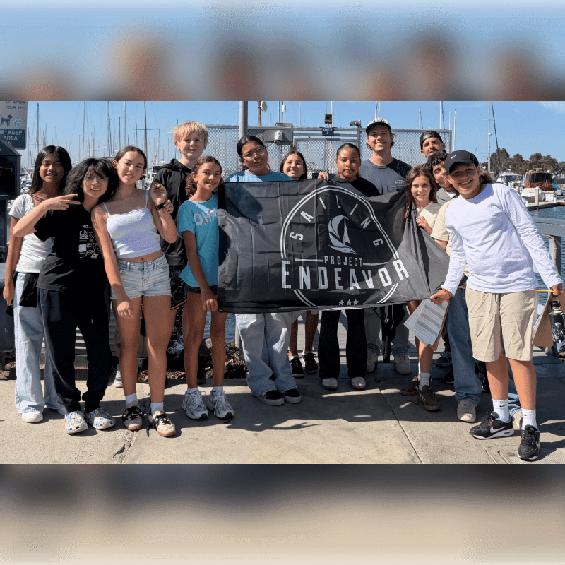 A group of young people hold a "Project Endeavor" sailing banner in the foreground, with a harbor and sailboats in the background.
