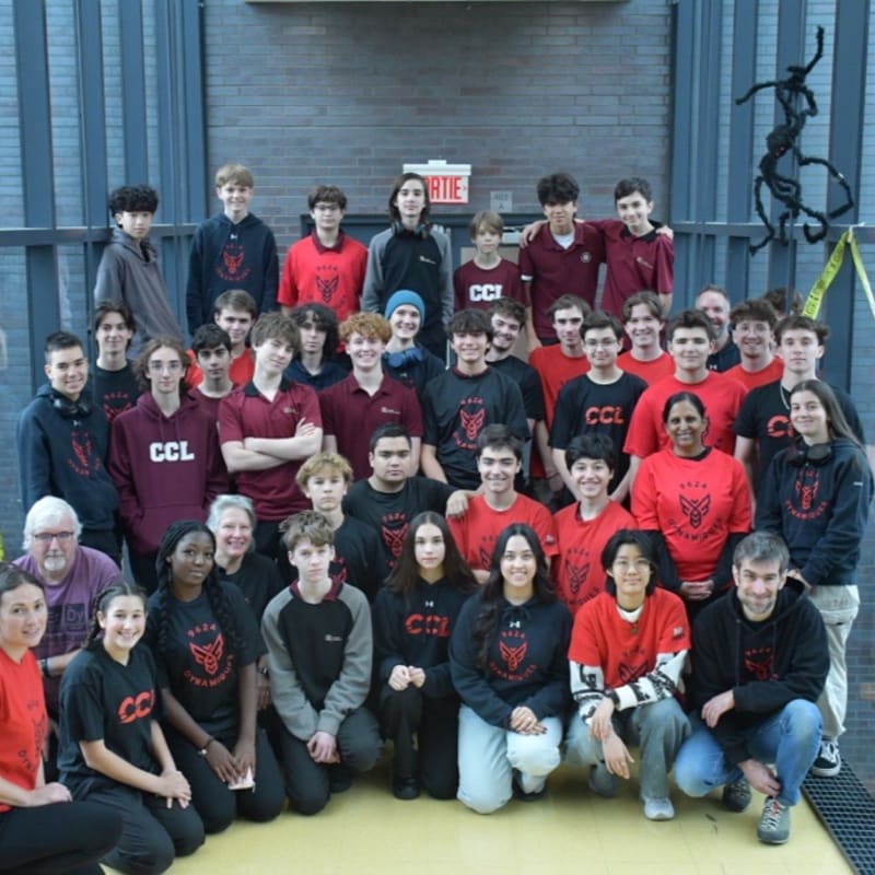 A large group of students in red and black clothing pose for a photo in front of a grey brick wall and metal doors.