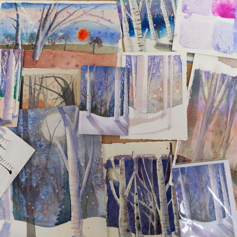 A collection of winter-themed watercolor paintings featuring birch trees and moons is scattered across a light-colored surface.