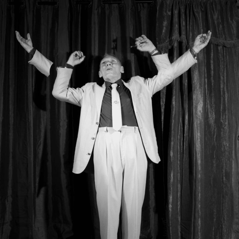 A man in a light-colored suit and tie stands with his arms raised in front of dark, heavy curtains.