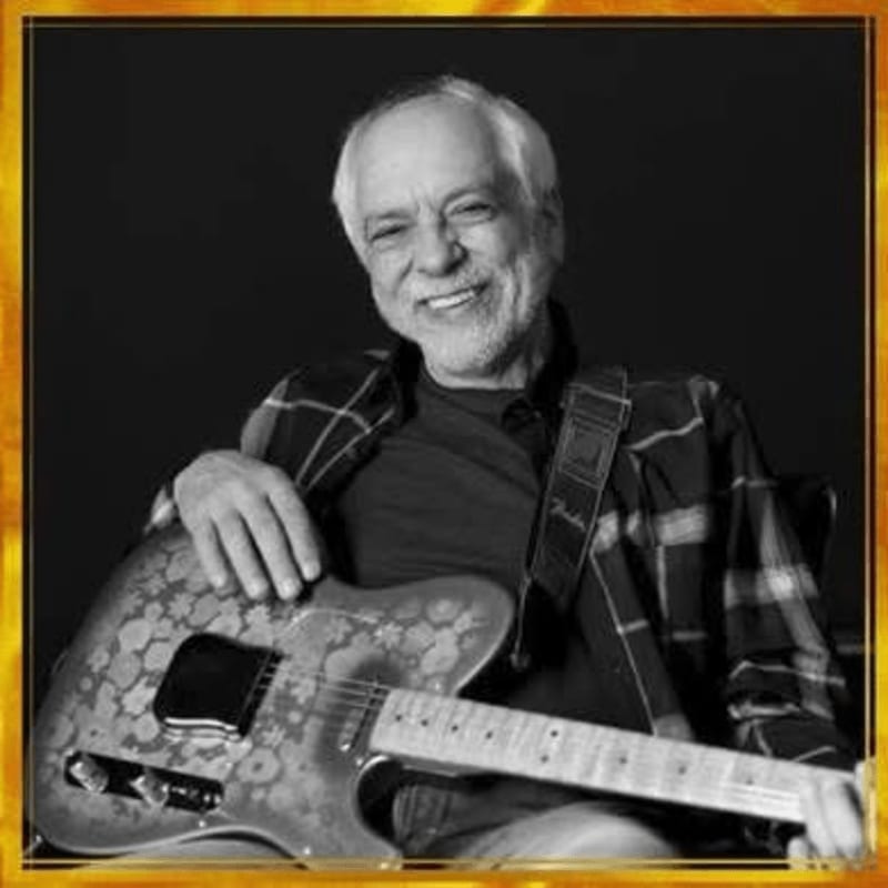 A smiling man in a plaid shirt holds an electric guitar against a solid black background.