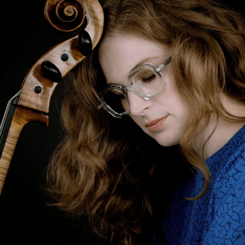A woman with curly red hair and clear glasses rests her head against the scroll of a cello against a solid black background.