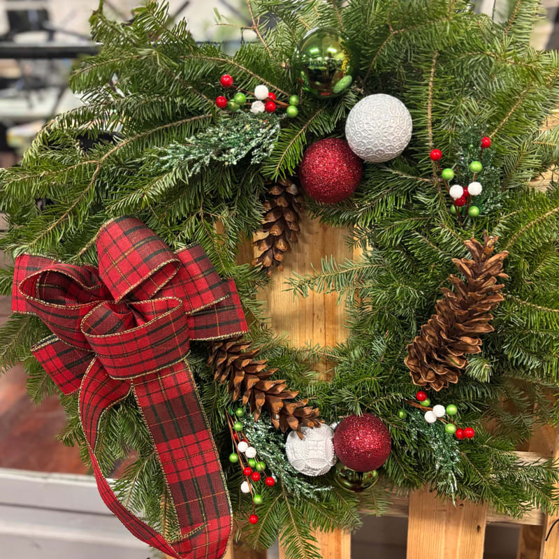 A festive evergreen wreath decorated with red and white ornaments, pinecones, and a large red plaid bow rests against a light-colored wooden background.