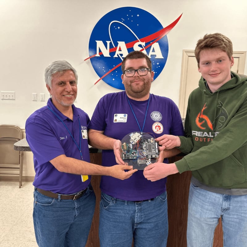 Three men stand together holding a circular electronic device in the foreground, with a large NASA logo mounted on the wall in the background.