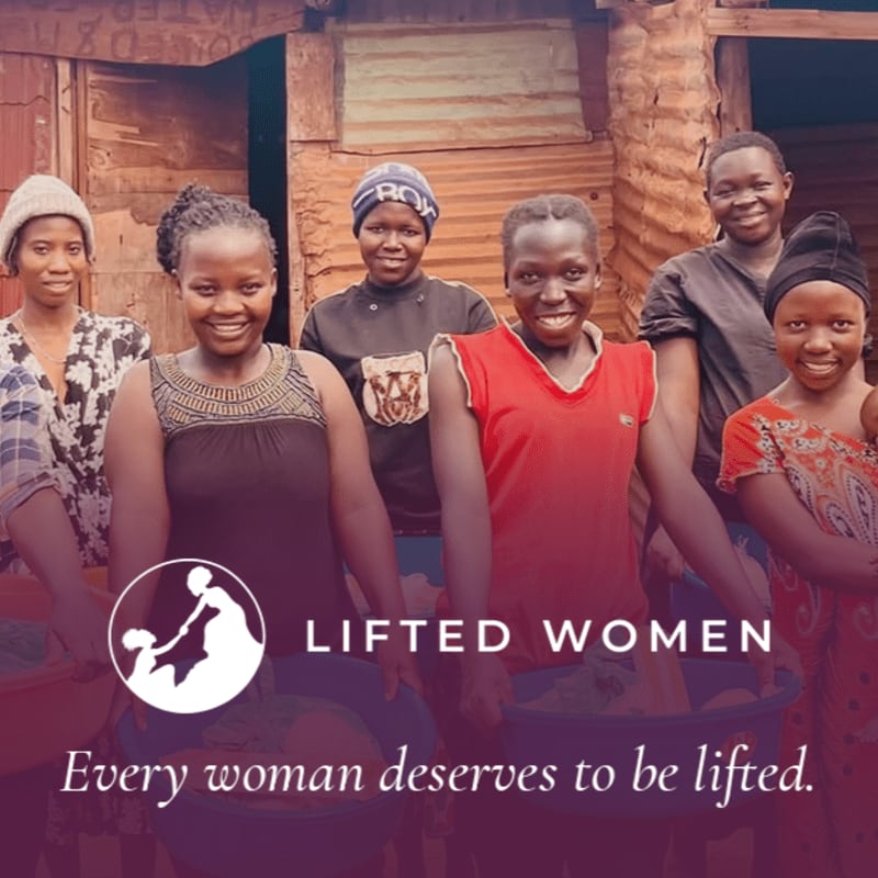 Six smiling women stand together in the foreground with basins, set against a rustic wooden building background.