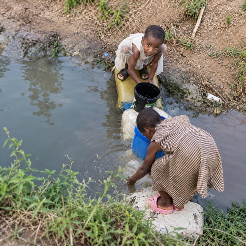 Two young children collect water from a murky stream using buckets, surrounded by dry earth and green foliage.