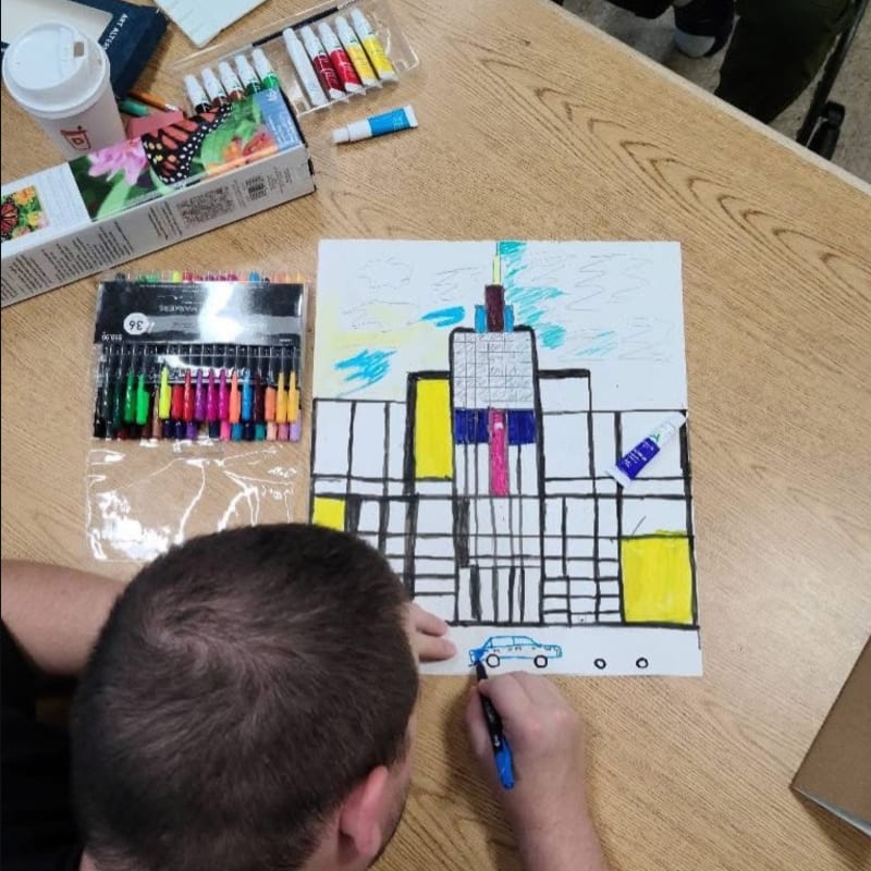 A person draws a blue car on a city skyline picture, surrounded by markers and art supplies on a wooden table.