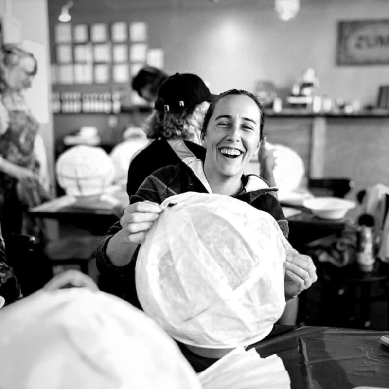 A laughing woman holds a large, white, wrapped object in the foreground of a busy workshop with other people and equipment in the background.