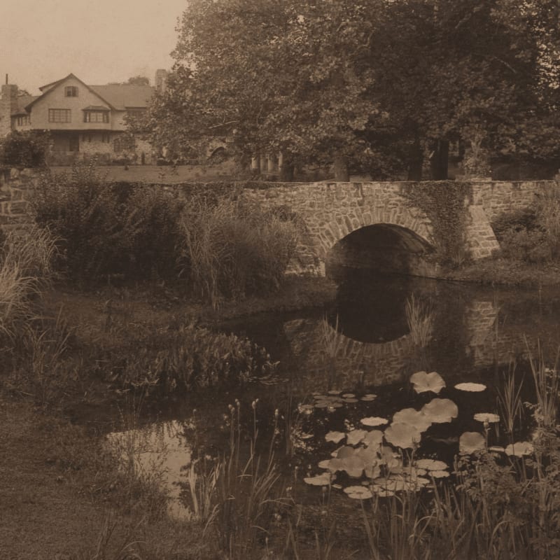 Water lilies float in a pond in the foreground, leading to a stone arch bridge and a house nestled among trees in the background.