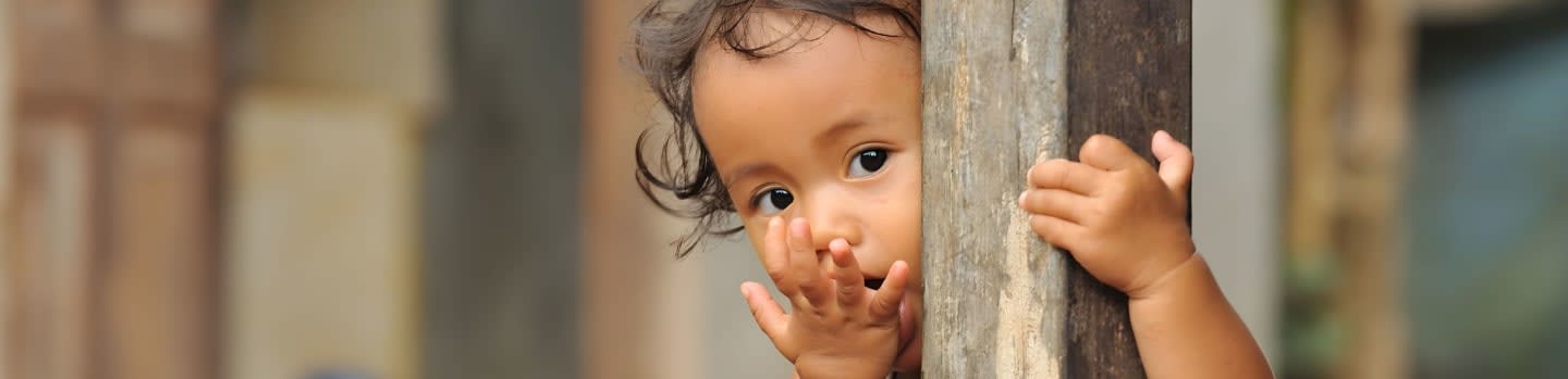 A young child peeks around a wooden post with a blurred background of a building.