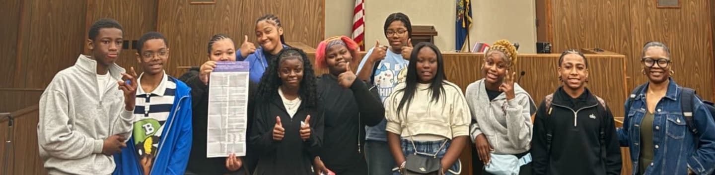 A group of smiling teenagers poses in a courtroom with a judge's bench and American flag in the background.