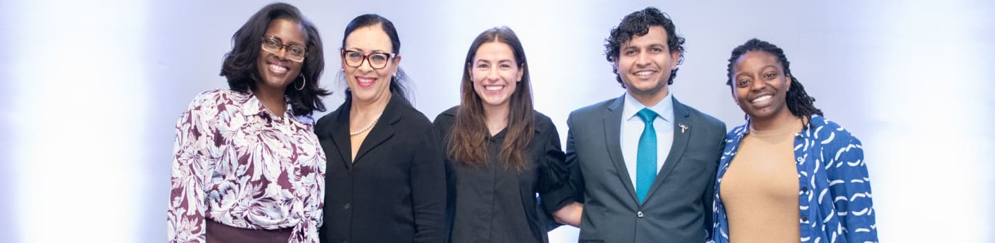 Five smiling individuals stand in front of a softly lit, pale blue background.