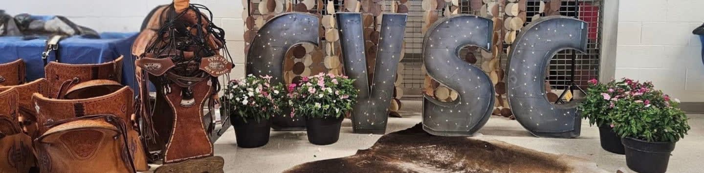 Several leather saddles are in the foreground, with large, textured letters spelling "GISC" and potted plants in the background.