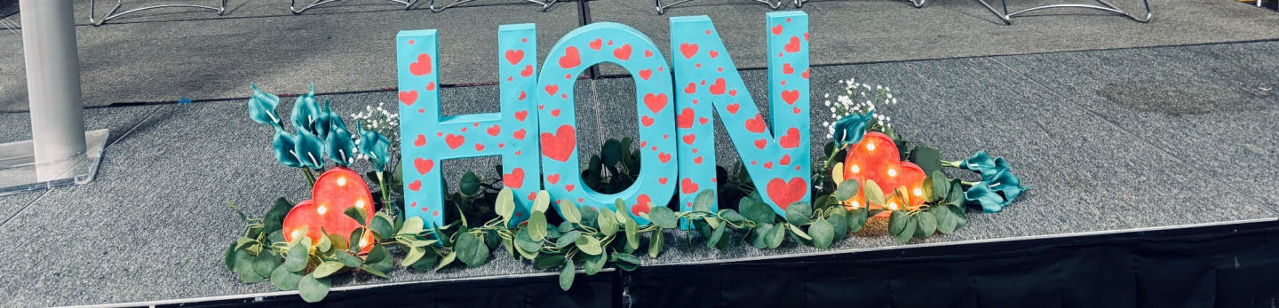 Large blue letters spelling "HON" decorated with hearts are displayed on a bed of green leaves and orange heart-shaped decorations, with a gray, textured surface in the background.