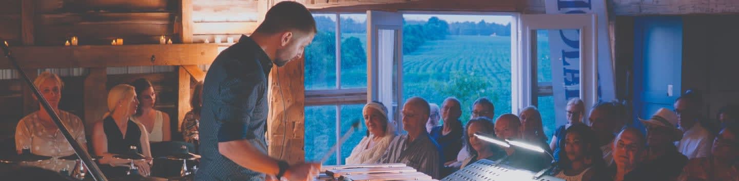 A man in a dark shirt addresses an audience seated in a rustic barn, with a field visible through large windows in the background.