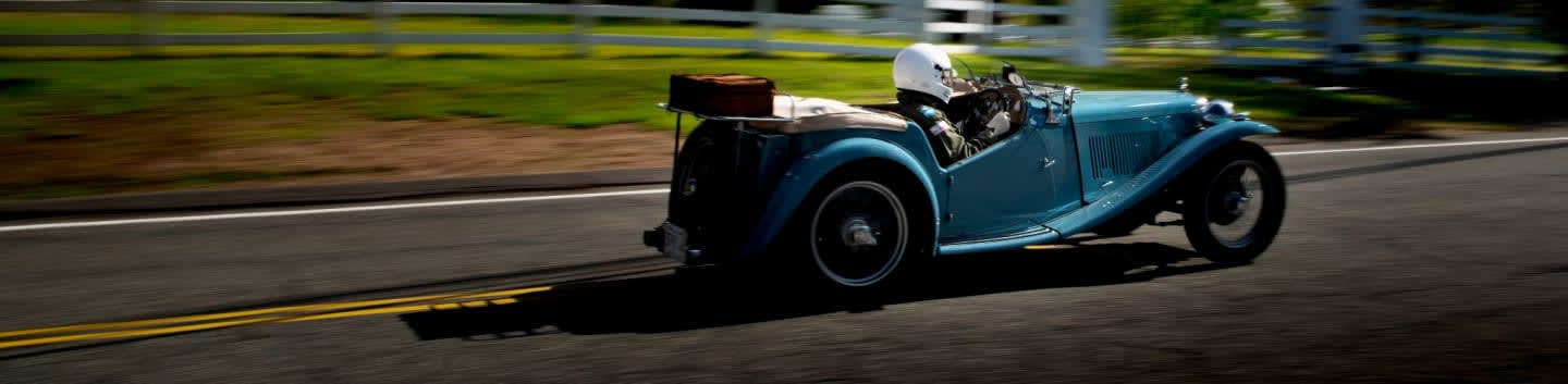 A blue vintage convertible speeds down a rural road with a white fence and trees blurred in the background.