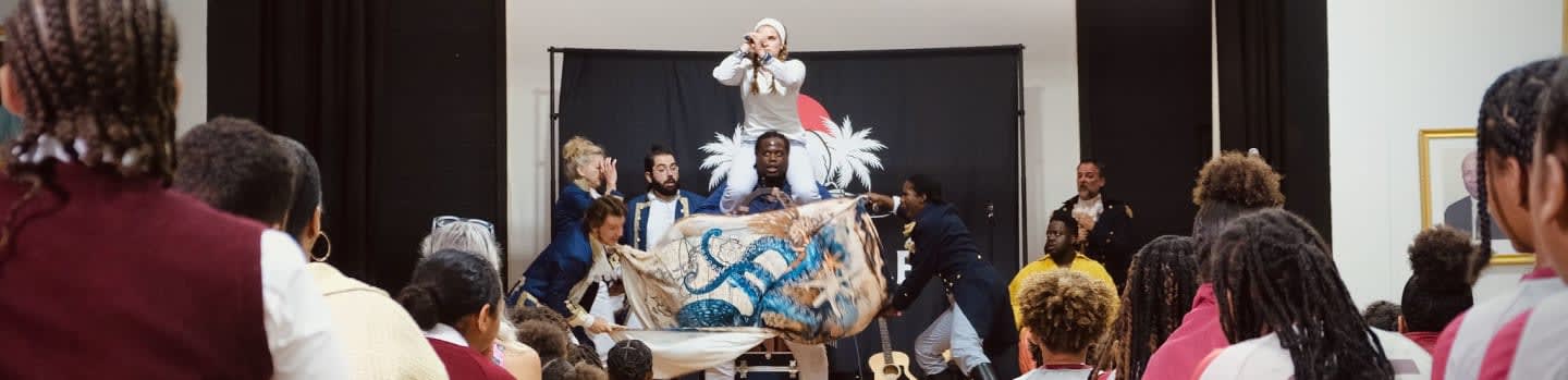 In the foreground, students in maroon and white uniforms watch a performance, while on the background stage, performers present a dramatic scene with a banner and a person standing on a raised platform.