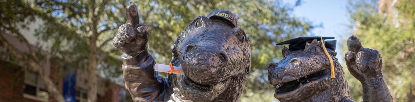 Two bronze alligator statues, one holding a diploma and the other wearing a graduation cap, stand in front of a blurred background of trees and buildings.