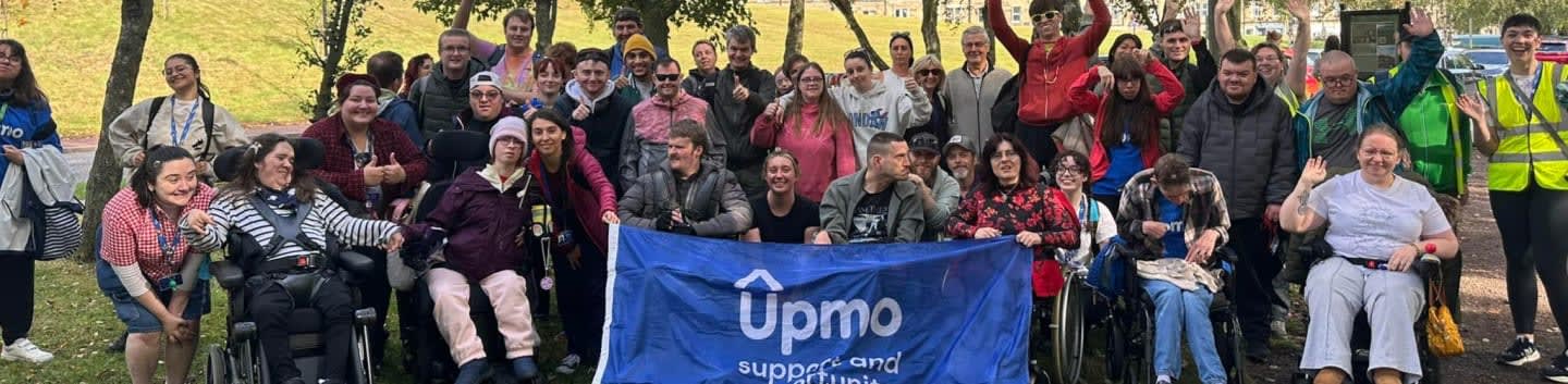 A large group of people, some in wheelchairs, pose for a photo in front of a blue banner with the text "Upmo support and" in a park setting with trees in the background.