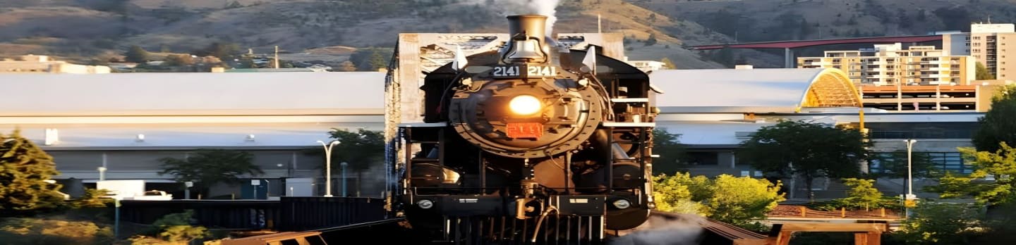 A vintage steam train with a lit headlight moves forward past modern buildings and a hillside in the background.