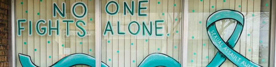 A teal ribbon and the words "NO ONE FIGHTS ALONE" are painted on a window with a light wood-paneled background.