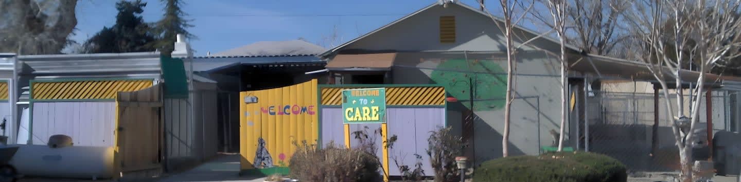 A yellow gate with "Welcome to Care" written on it stands in front of a building with a corrugated metal roof and pastel-colored walls, while bare trees and bushes are visible in the background.