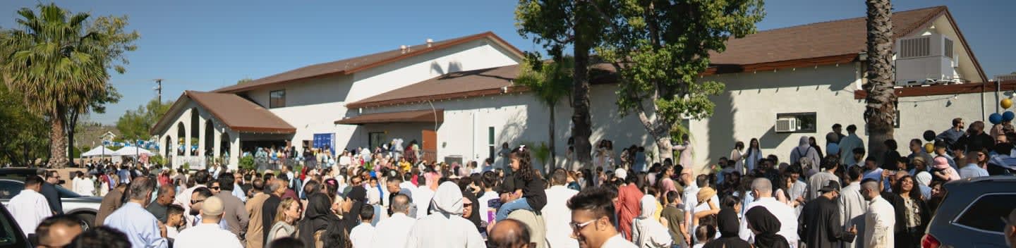 A large crowd of people gathers in front of a building with a brown roof under a clear blue sky.