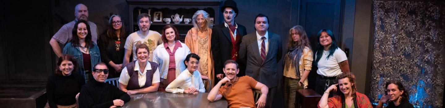 A group of people in casual and formal attire pose in front of a dimly lit stage set with shelves and a textured wall.