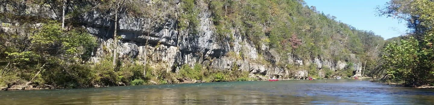 A calm river flows in the foreground, reflecting the rocky, tree-covered cliffs that rise steeply in the background under a clear blue sky.