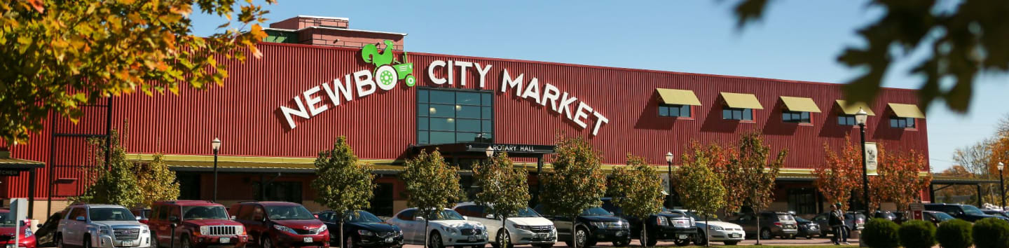 Cars are parked in front of the NewBo City Market building, with autumn foliage visible in the foreground and background.