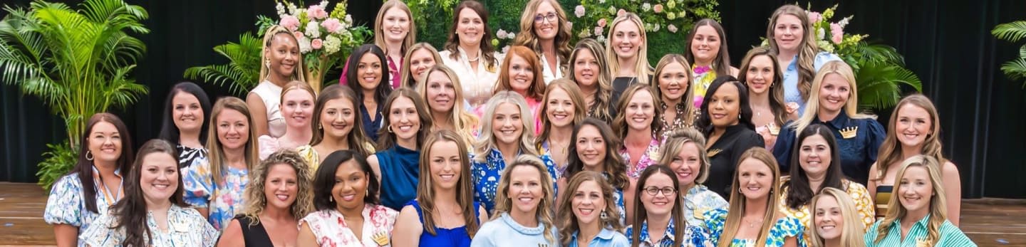 A large group of women smile for a photo in front of lush green foliage and a dark backdrop.