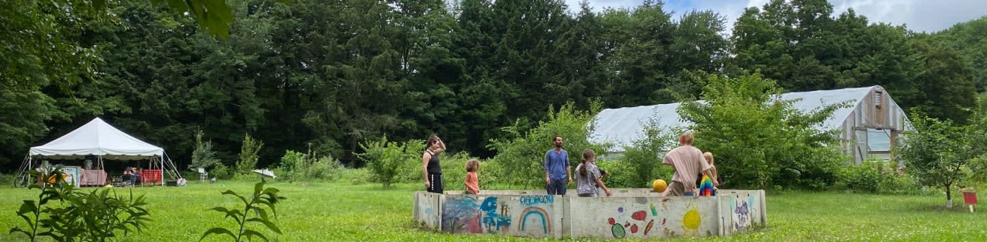A group of people are gathered around a decorated concrete structure in a grassy field, with a white tent and a greenhouse visible in the background among the trees.