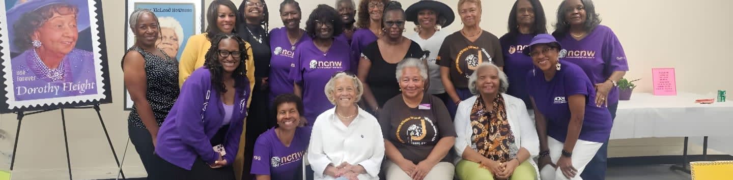 A group of women in purple shirts pose in front of a display featuring a stamp of Dorothy Height, with a table in the background.