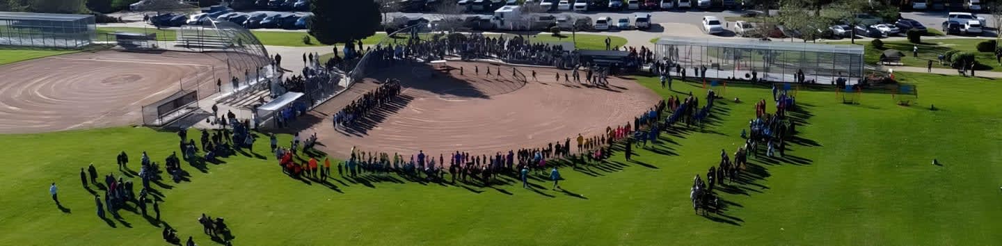 A large group of people are gathered in a circle on a baseball field, with a parking lot and trees in the background.