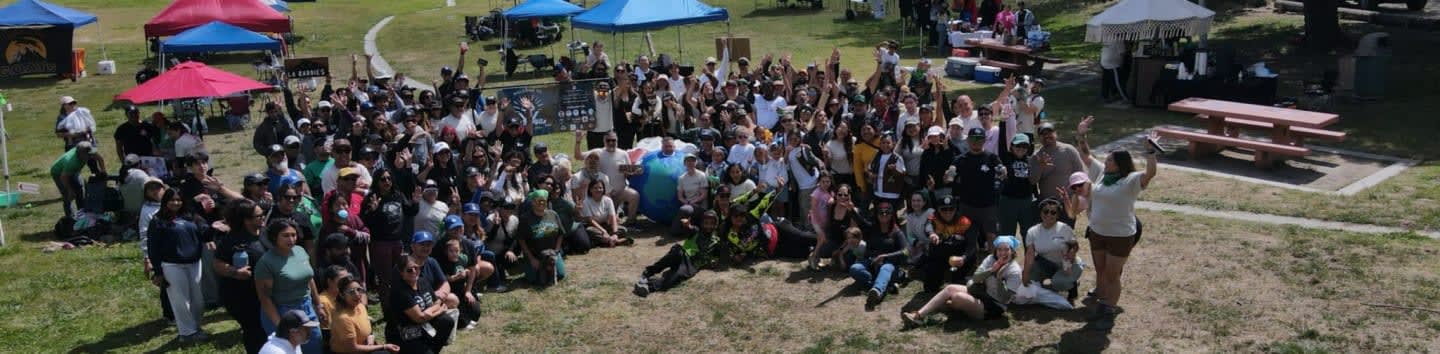 A large group of people are gathered on a grassy field in front of tents and picnic tables.