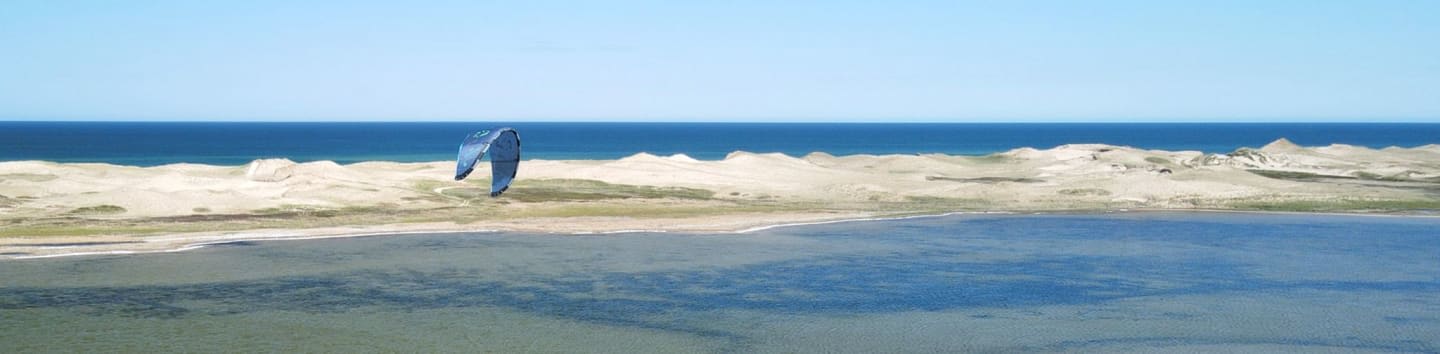 A blue kite soars over a sandy beach with calm water in the foreground and the ocean in the background.