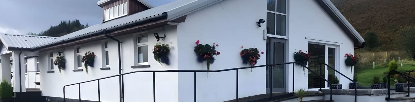 A white building with hanging flower baskets is set against a backdrop of a tree-covered hillside.