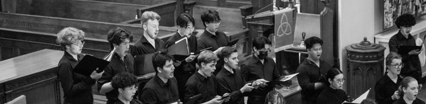 A choir of young people in black robes sings from sheet music in front of wooden pews and a decorative banner.
