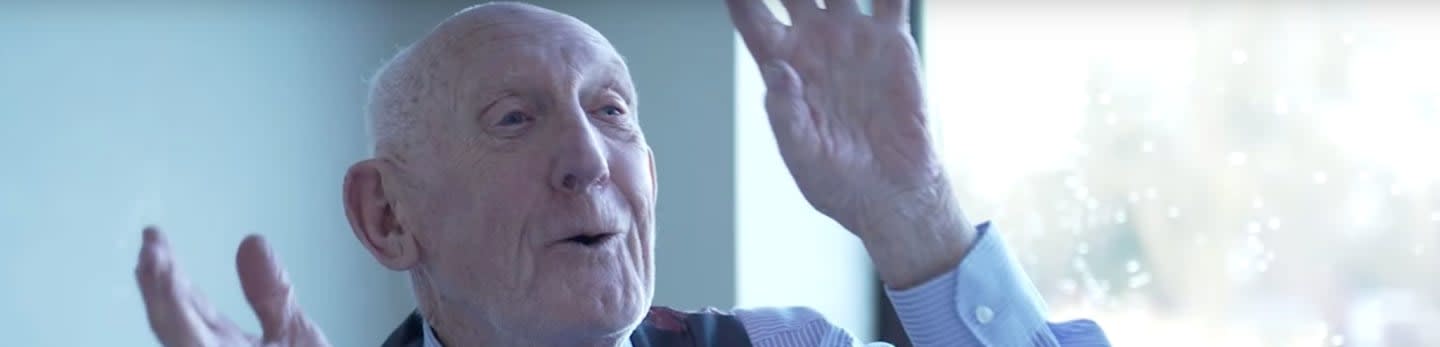 An elderly man with his hands raised in a gesture of speech is in the foreground, with a bright, out-of-focus background.
