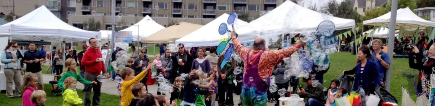 A clown blows bubbles for a crowd of people gathered at an outdoor event with tents and buildings in the background.