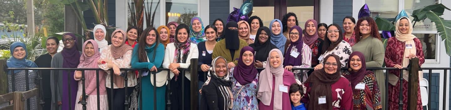 A group of women in hijabs pose for a photo in front of a building with a railing in the foreground.