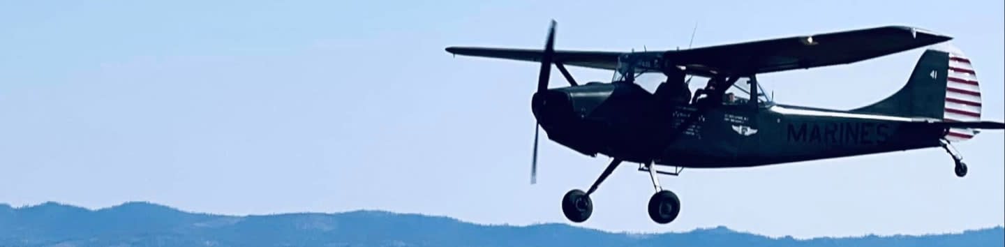 A dark-colored biplane flies over a hazy blue mountain range against a clear sky.