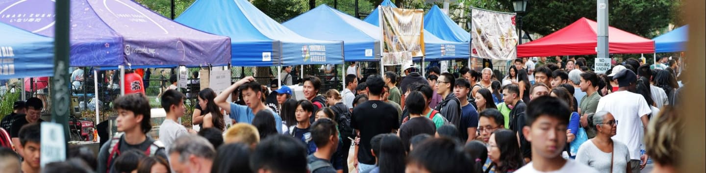 A crowd of people mills about in front of colorful tents at an outdoor market.