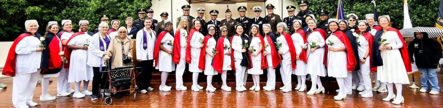 A group of people in uniform, including some in white outfits with red capes and others in military attire, pose for a photo in front of a building with a tree-lined background.