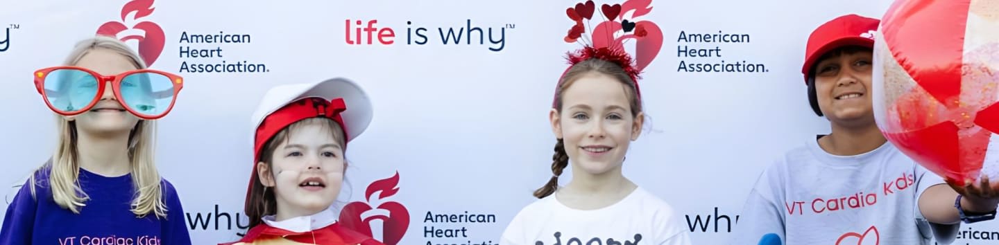 Four children, two wearing oversized glasses and hats, pose in front of a white backdrop with "American Heart Association" and "life is why" logos.