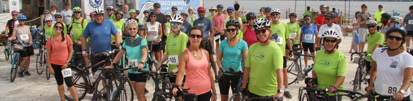 A group of cyclists in bright shirts and helmets pose with their bikes in the foreground, while a blurred background shows more participants and a building.
